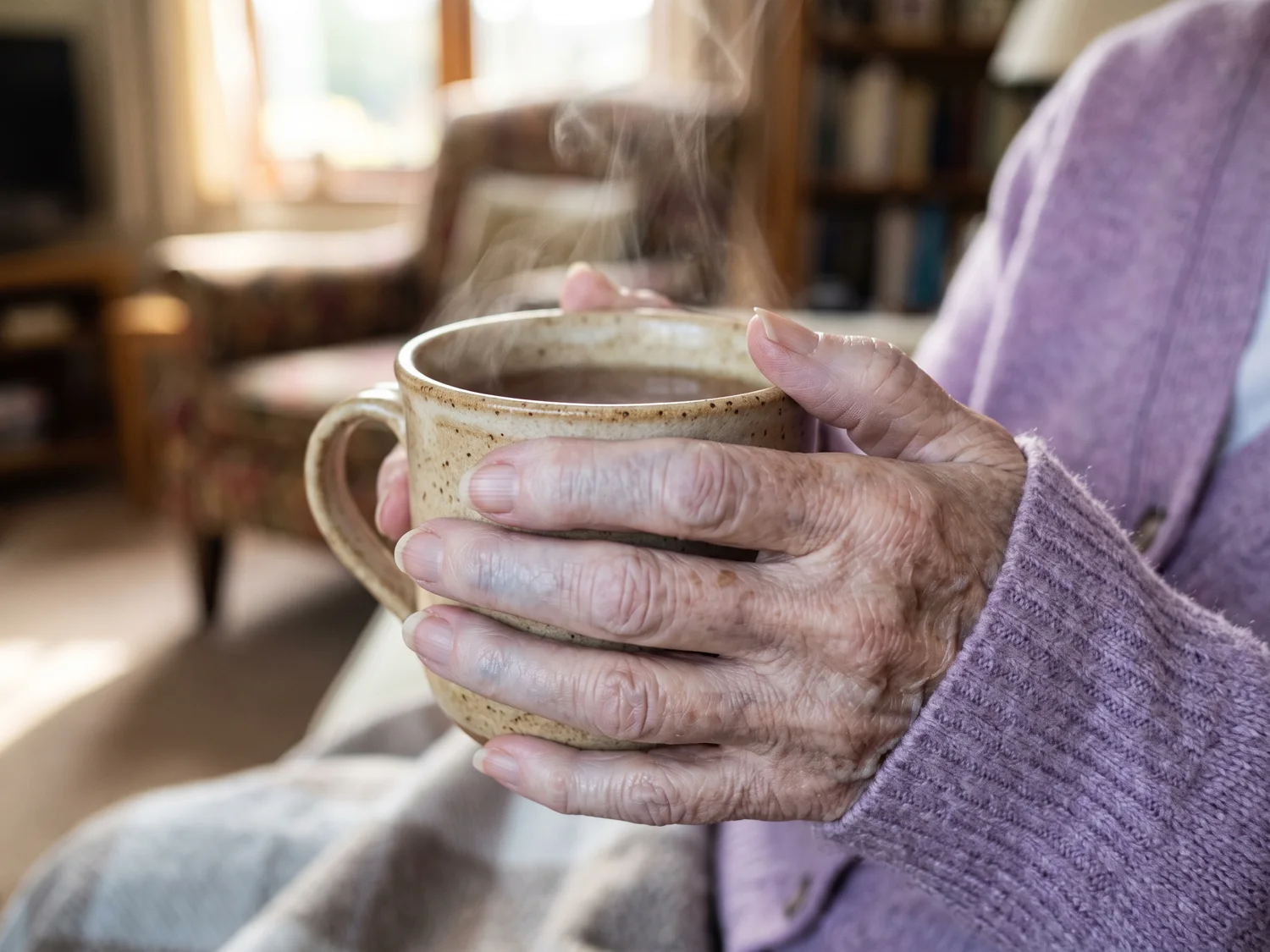 Close-up of an elderly woman's pale cold fingers wrapped around a warm mug in a sunlit living room
